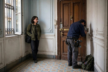 Técnico cerrajero con cinturón de herramientas examinando y manipulando con precisión la cerradura de una puerta señorial antigua en un rellano de mosaico hidráulico mientras la clienta observa con calma.