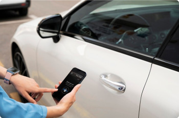Propietario utilizando una interfaz digital móvil para la apertura remota de un vehículo blanco de gama alta, representando la convergencia tecnológica en seguridad vial.
